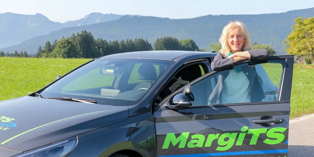 Lächelnde Fahrlehrerin steht neben ihrem Auto mit Berglandschaft im Hintergrund.